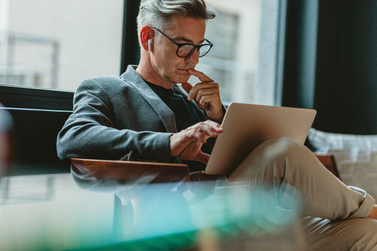 Ein Mann mit grauem Haar und Brille sitzt auf einem Sessel und arbeitet konzentriert an einem Laptop. Er trägt einen dunkelgrauen Blazer und denkt nach, während er seine Hand an das Kinn legt. Hinter ihm sind große Fenster in einem modernen Raum zu sehen.