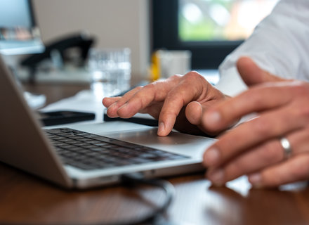Arbeiten am Laptop im Büro Hände eines Mannes, die auf einer Laptop-Tastatur tippen. Im Hintergrund sind ein Glas Wasser und ein Smartphone sichtbar. Der Arbeitsplatz ist hell und modern eingerichtet.