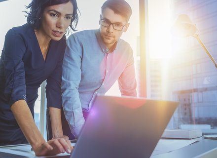 Zwei Personen stehen an einem Tisch und schauen auf einen Laptop. Eine Frau mit dunklem Haar trägt ein schwarzes Kleid und zeigt auf den Bildschirm, während ein Mann mit Brille und Hemd interessiert zusieht. Im Hintergrund sind Fenster mit Stadtansicht zu sehen.