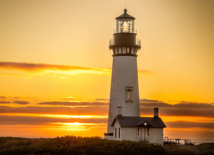 Ein Leuchtturm steht auf einer Klippe bei Sonnenuntergang, umgeben von sanften Hügeln. Die warme Farbpalette des Himmels reicht von Gelb über Orange bis zu einem sanften Blau, während das Licht des Leuchtturms sichtbar ist.