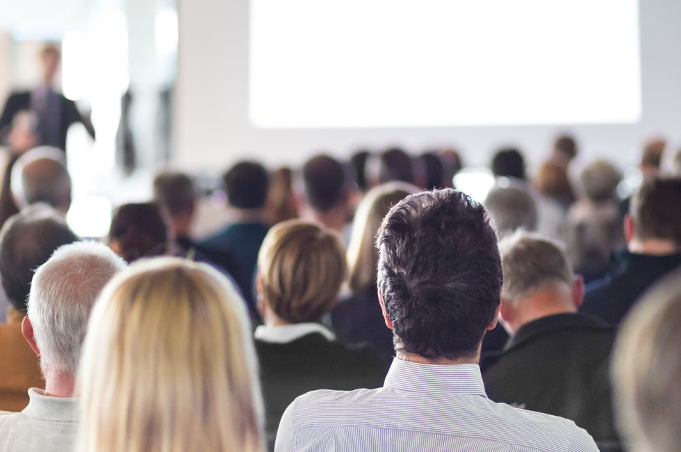 Eine Gruppe von Personen sitzt in einem Seminarraum und schaut auf eine Präsentation. Im Vordergrund sind die Rücken der Teilnehmer sichtbar, während im Hintergrund ein Redner steht und spricht.